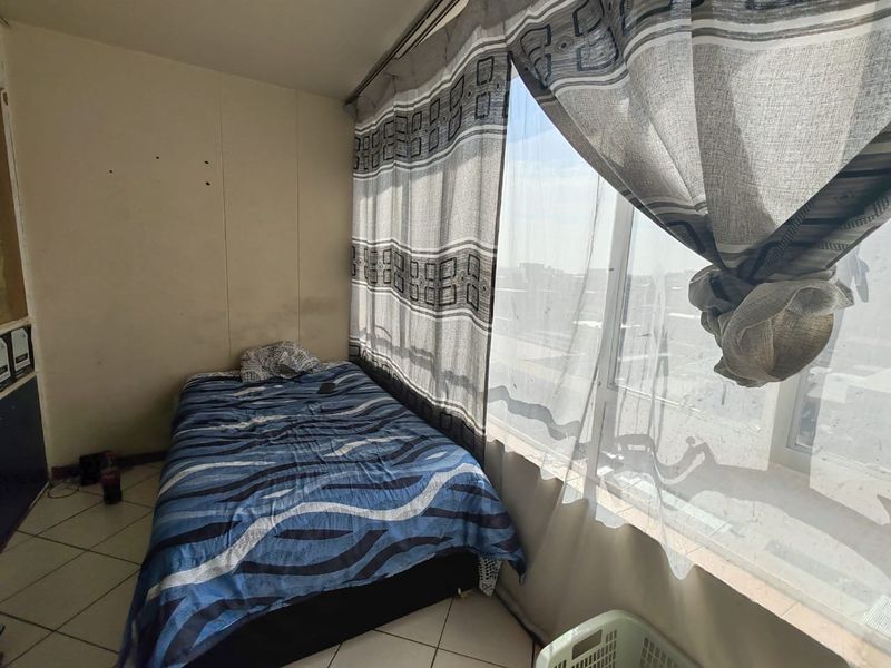 A compact bedroom featuring tiled flooring and a large window that provides ample natural light, currently requiring minor cosmetic wall repairs.