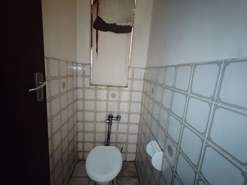 A compact, dated bathroom featuring ceramic wall and floor tiling, a flush toilet, and a window with a damaged covering.