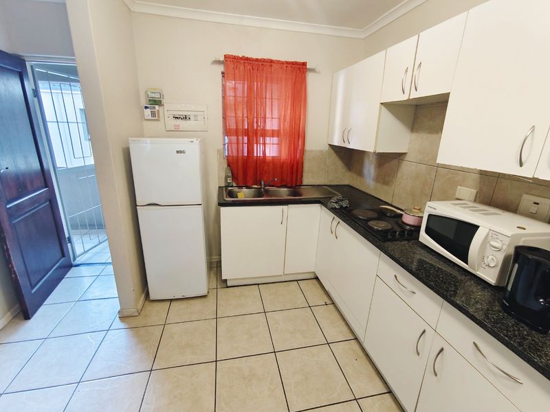 Compact kitchen featuring white built-in cabinetry, dark granite countertops, a double basin sink, electric hob, and tiled flooring with direct access to an exterior area.