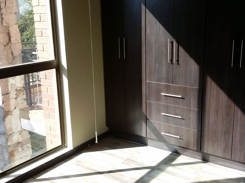A bright bedroom featuring dark wood built-in cupboards, tiled flooring, and a large window with views of the exterior brick facade.