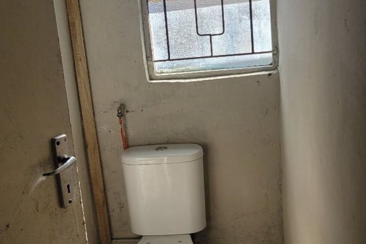 A small, basic bathroom featuring a standard toilet, tiled flooring, and a window with security bars, currently requiring cosmetic renovation and wall repairs.