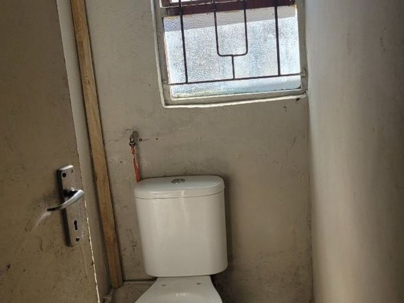 A small, basic bathroom featuring a standard toilet, tiled flooring, and a window with security bars, currently requiring cosmetic renovation and wall repairs.