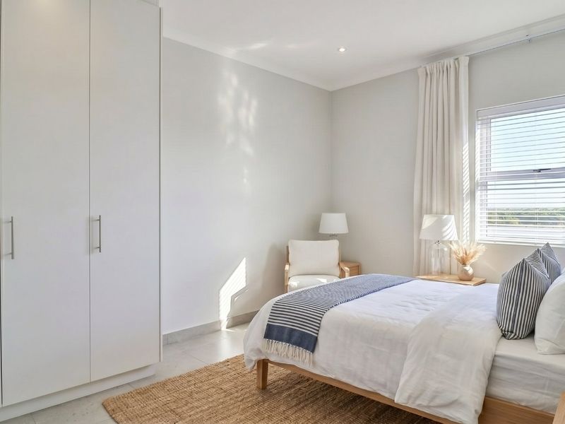 A bright and modern bedroom featuring neutral tones, tiled flooring, built-in wardrobe storage, and natural light through a window with venetian blinds.