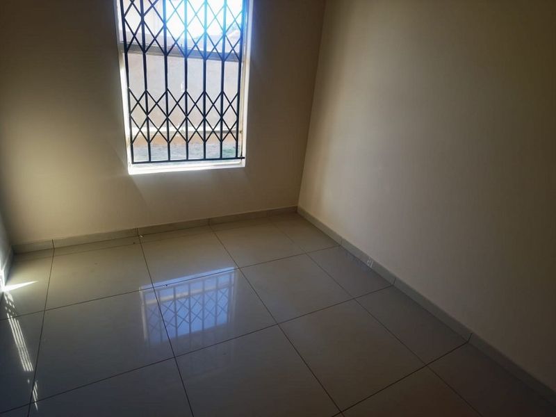 A bright, empty bedroom featuring neutral tiled flooring and a window fitted with a security gate.