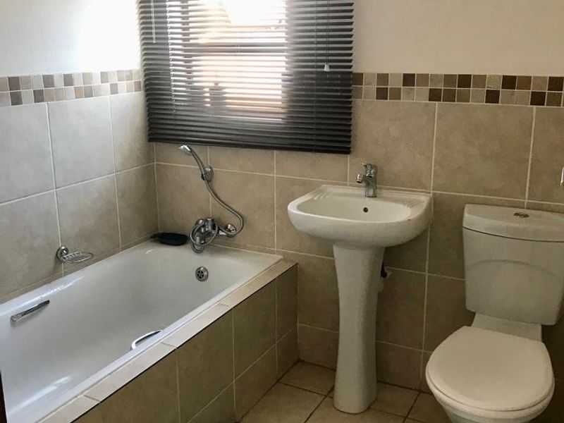 A functional bathroom featuring a pedestal basin, built-in bathtub, and toilet with neutral wall tiling and a window with a venetian blind.