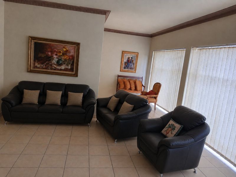 A spacious living room featuring neutral ceramic floor tiles, crown molding, and large windows fitted with vertical blinds.