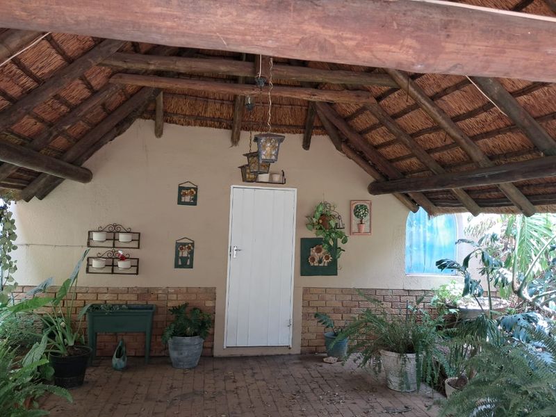 A rustic covered patio area featuring a traditional thatched roof with exposed timber beams, brick base walls, and paved flooring.