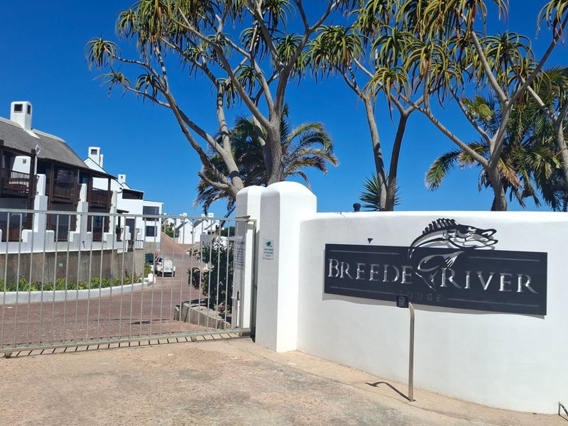 Entrance view of Breede River Lodge featuring a secure gated access point, paved driveway, and white exterior buildings with traditional thatched roofing.