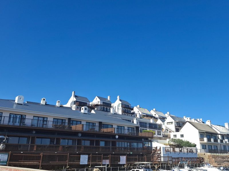 A view of a multi-level apartment complex featuring white facades, metal roofing, and timber-railed balconies overlooking a waterfront area.