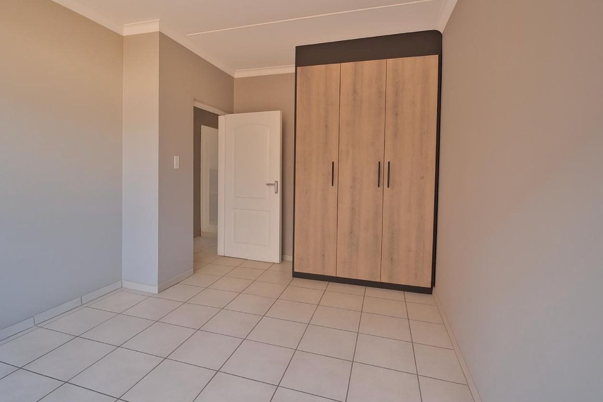 A clean, empty bedroom featuring neutral wall tones, light-colored floor tiles, and a modern built-in wardrobe with wood-grain finish.
