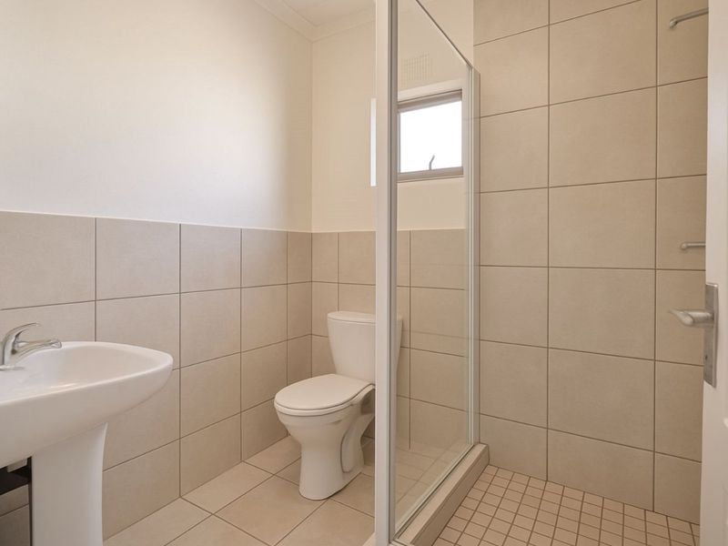 A clean, functional bathroom featuring a pedestal basin, toilet, and a glass-enclosed shower with neutral wall and floor tiling.