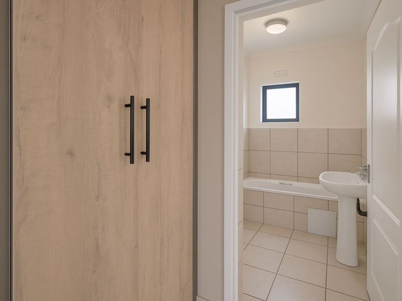 A clean, neutral bathroom featuring a pedestal basin, built-in bathtub, and tiled finishes, adjacent to a hallway with a built-in cupboard.