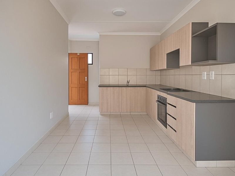 A clean, modern kitchen featuring light wood-tone cabinetry, dark countertops, built-in oven and hob, tiled backsplash, and neutral floor tiles.