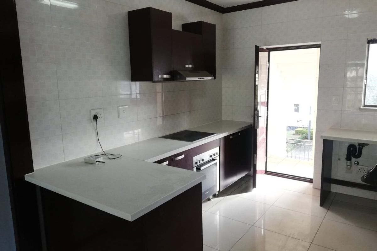 A clean, functional kitchen featuring dark cabinetry, stone countertops, built-in oven, electric hob, and tiled finishes with direct access to an outdoor area.