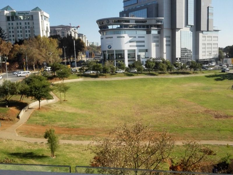 Elevated urban view overlooking a landscaped park area with surrounding commercial high-rise buildings and city infrastructure.
