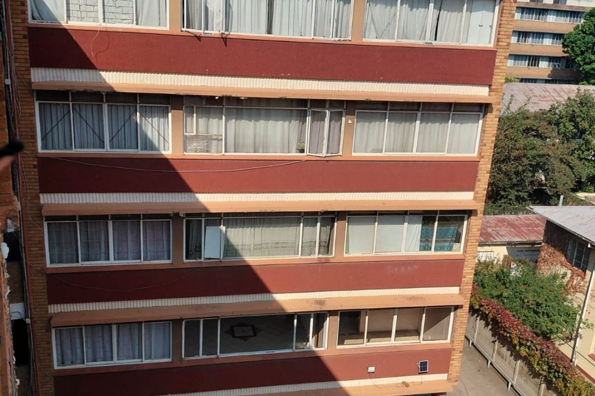 Rear elevation of a multi-story apartment building featuring brick facade, concrete support pillars, and a paved ground-level parking area.