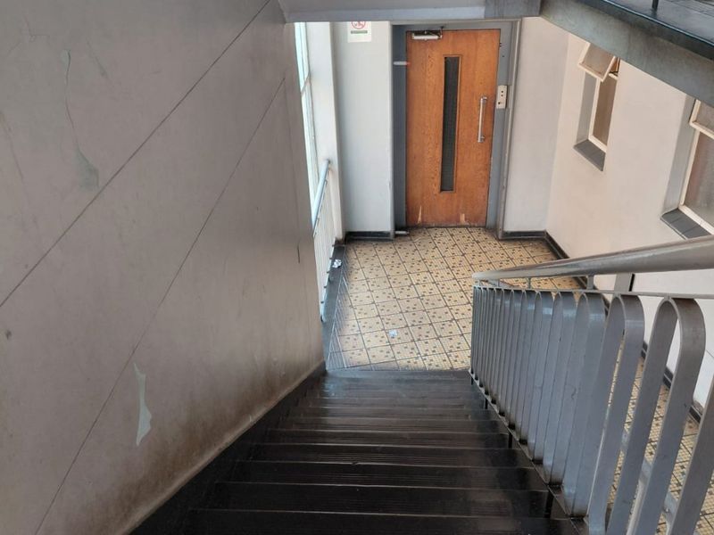 A communal apartment stairwell featuring metal railings, tiled flooring, and a wooden entrance door, showing signs of wear and peeling paint on the walls.