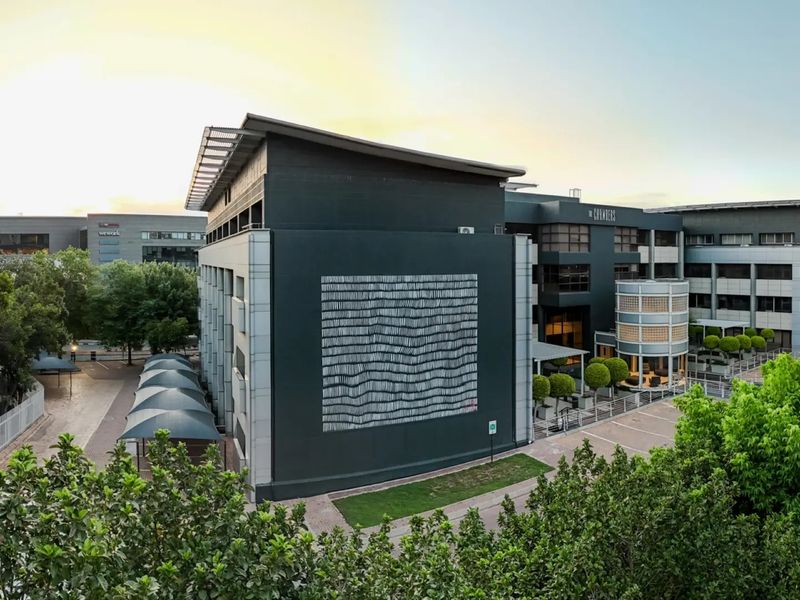A high-angle view of a modern commercial office complex featuring a dark facade, architectural mural, and covered parking facilities within a landscaped environment.