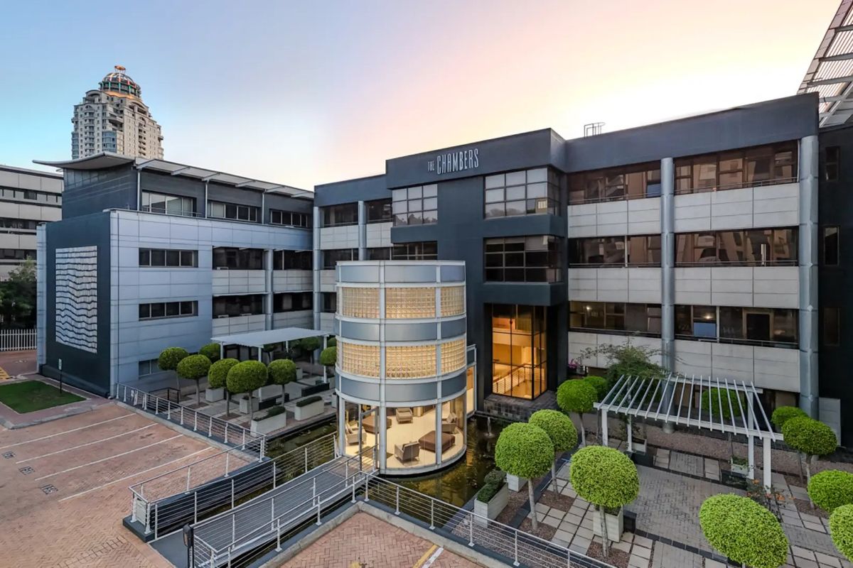 Modern commercial office building featuring a striking glass cylindrical entrance, water feature, pedestrian bridge, and landscaped courtyard with paved parking areas.