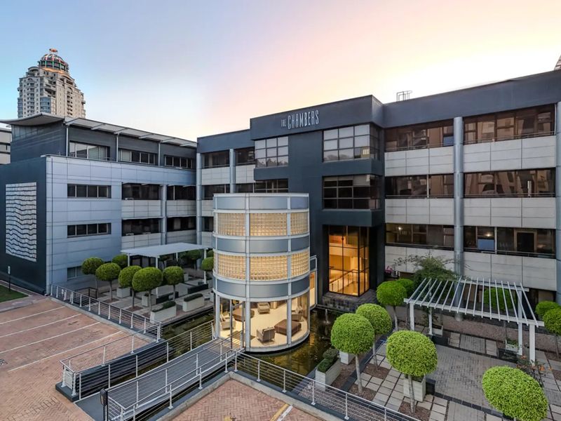 Modern commercial office building featuring a striking glass cylindrical entrance, water feature, pedestrian bridge, and landscaped courtyard with paved parking areas.