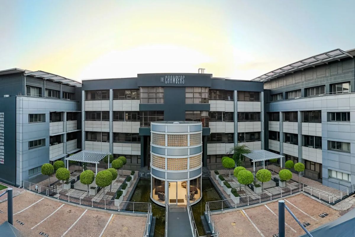 A professional panoramic view of The Chambers commercial office building, featuring a central glass block entrance, water feature, and paved parking area.