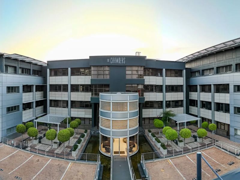 A professional panoramic view of The Chambers commercial office building, featuring a central glass block entrance, water feature, and paved parking area.