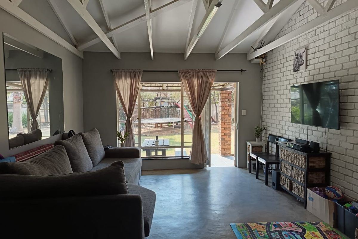 A bright living room featuring exposed timber roof trusses, a painted brick feature wall, polished concrete floors, and direct access to the rear garden.