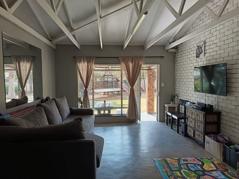 A bright living room featuring exposed timber roof trusses, a painted brick feature wall, polished concrete floors, and direct access to the rear garden.