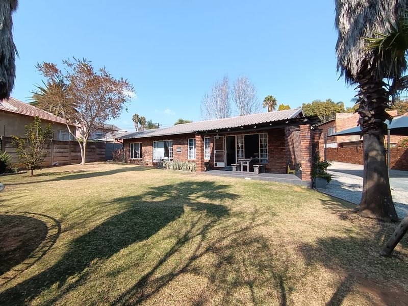 Rear view of a single-story brick house featuring a covered patio area, a spacious lawn, and established garden surroundings.