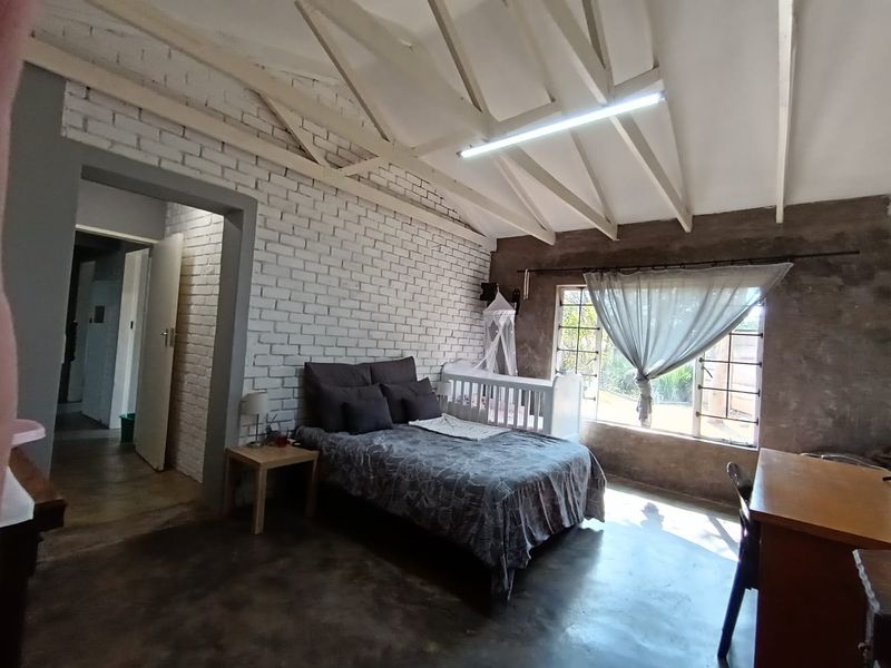 Spacious bedroom featuring exposed timber roof trusses, painted brick feature wall, and polished concrete flooring with large steel-framed windows.