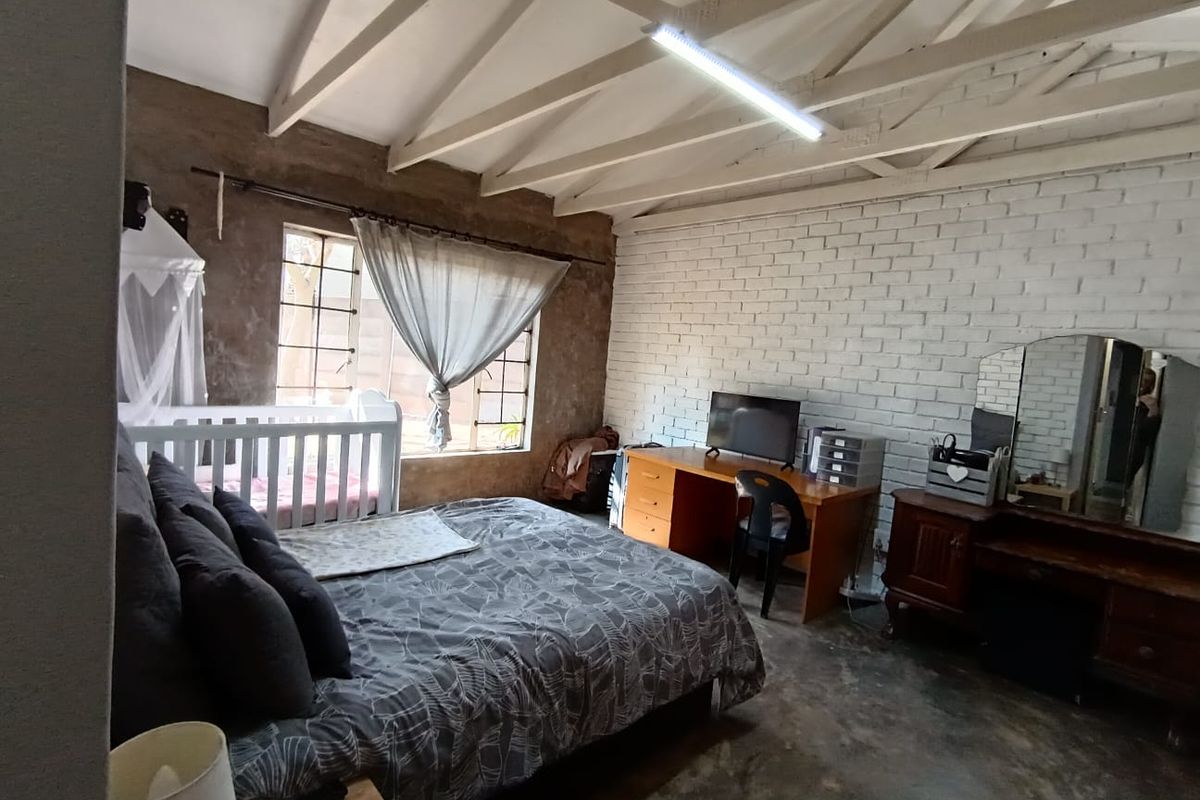 Spacious bedroom featuring exposed timber rafters, a painted brick feature wall, and industrial-style concrete flooring with large steel-framed windows.
