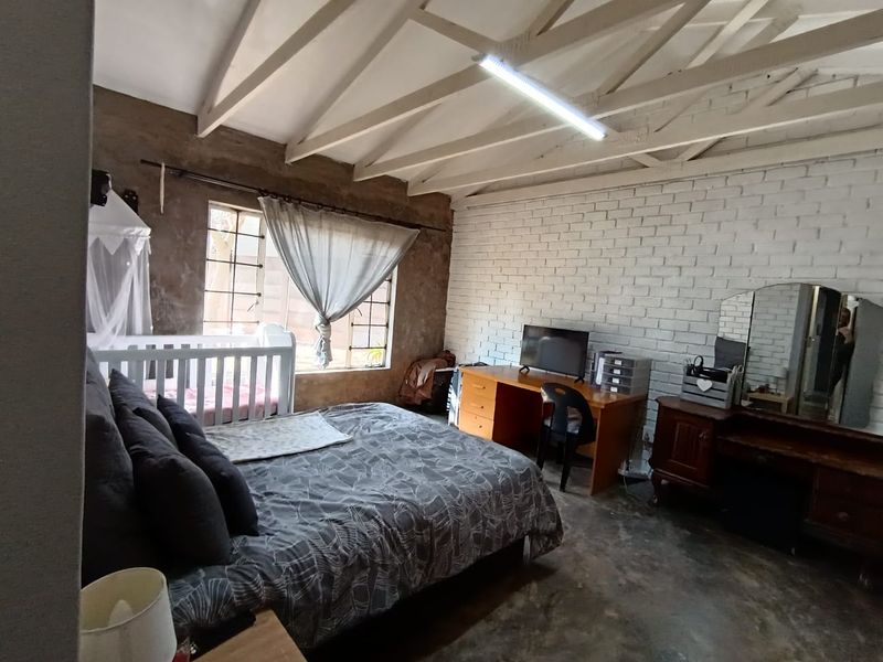Spacious bedroom featuring exposed timber rafters, a painted brick feature wall, and industrial-style concrete flooring with large steel-framed windows.