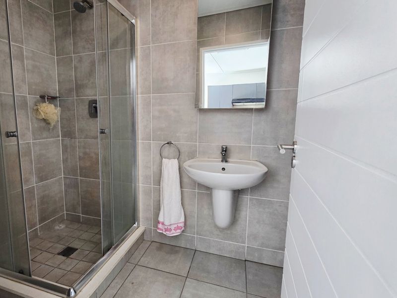 A modern bathroom featuring a glass-enclosed shower, pedestal washbasin, and neutral grey wall and floor tiles.