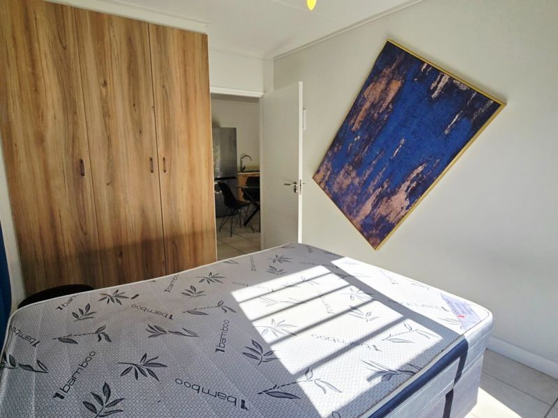 A bedroom featuring built-in wooden cupboards and tiled flooring, with a view through the doorway into the adjacent kitchen area.