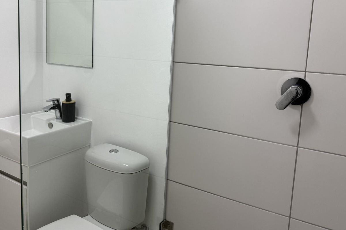 A modern, compact bathroom featuring a vanity basin, toilet, and a glass-enclosed shower with neutral wall tiling.