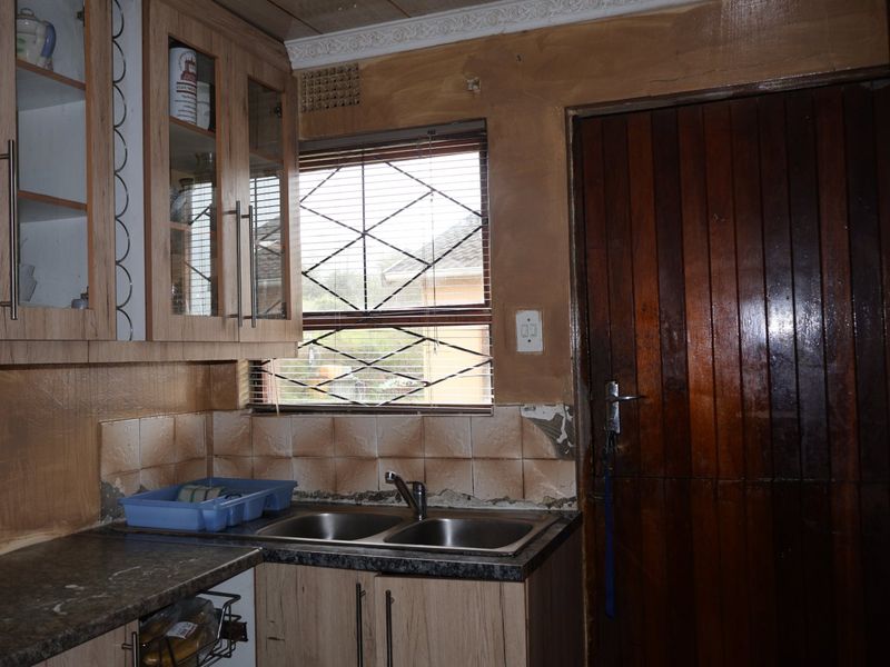 A kitchen area featuring laminate cabinetry, a double bowl stainless steel sink, and a timber door, requiring cosmetic updates to the wall finishes and tiling.