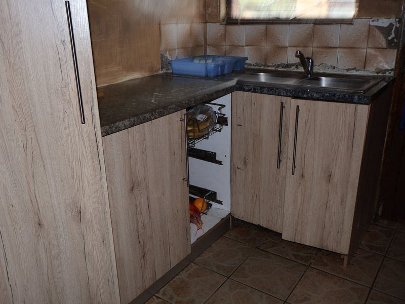 A kitchen area featuring wood-look cabinetry and a stainless steel sink, currently showing significant wear and damage to the wall tiles and cabinetry.