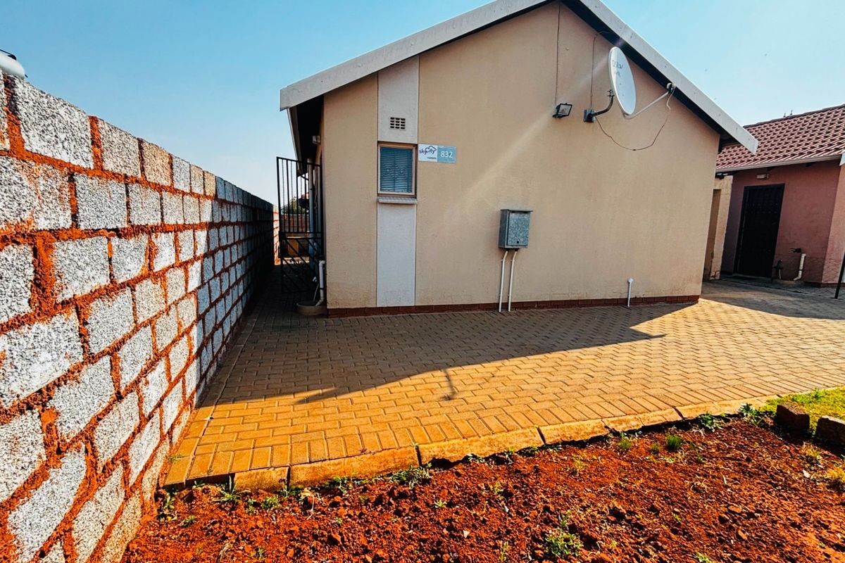 Rear exterior view of the property featuring a paved patio area, boundary wall, and external utility connections.