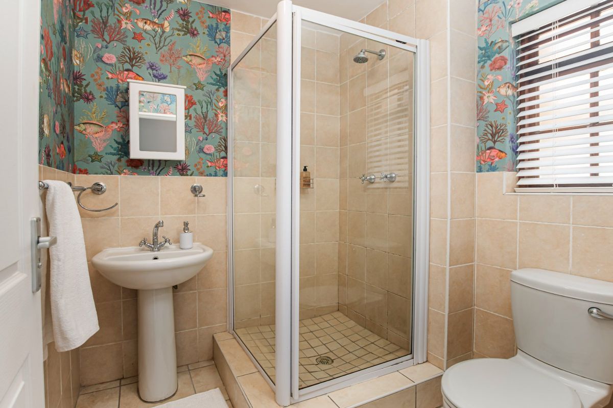 A functional bathroom featuring a pedestal basin, a glass-enclosed shower, and a toilet, finished with neutral wall tiles and decorative wallpaper.