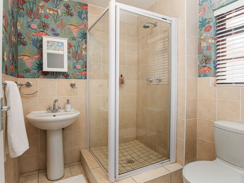A functional bathroom featuring a pedestal basin, a glass-enclosed shower, and a toilet, finished with neutral wall tiles and decorative wallpaper.