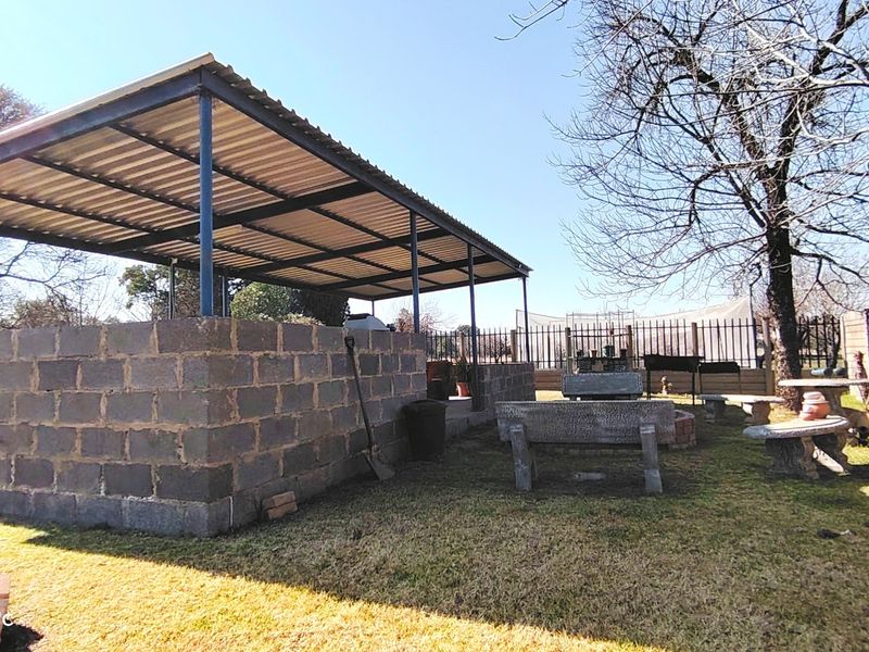 Outdoor covered entertainment area featuring a concrete block structure, corrugated metal roof, and fixed concrete seating in a garden setting.