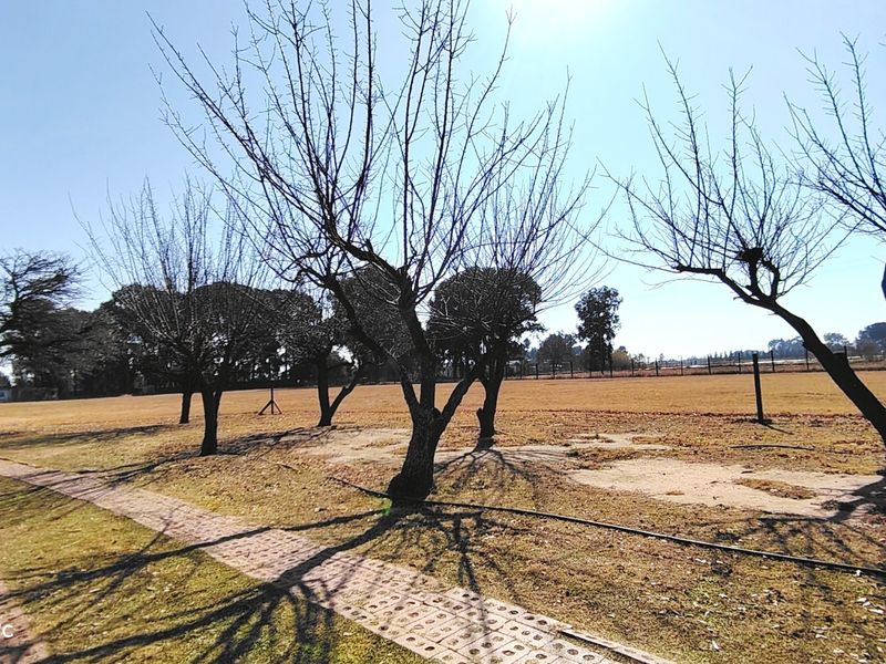A wide-angle view of a large, open farm garden area featuring mature trees, a paved walkway, and perimeter fencing under a clear blue sky.