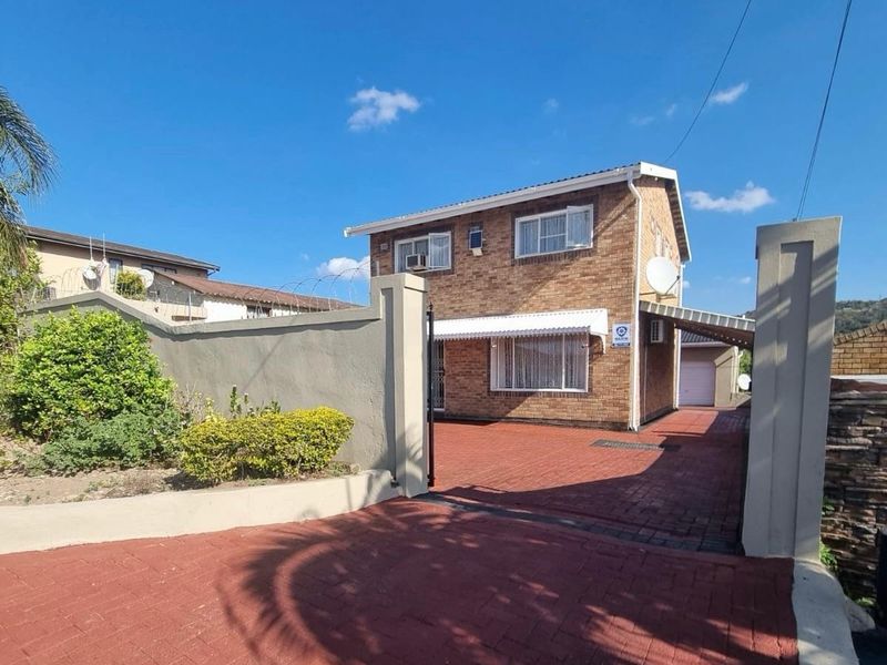 A well-maintained double-story face-brick home featuring a paved driveway, secure perimeter walling, and a convenient carport leading to a garage.