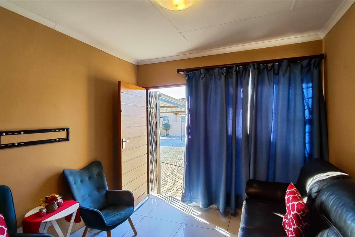 A living room area featuring tiled flooring, a timber entrance door with a security gate, and access to an outdoor paved area.