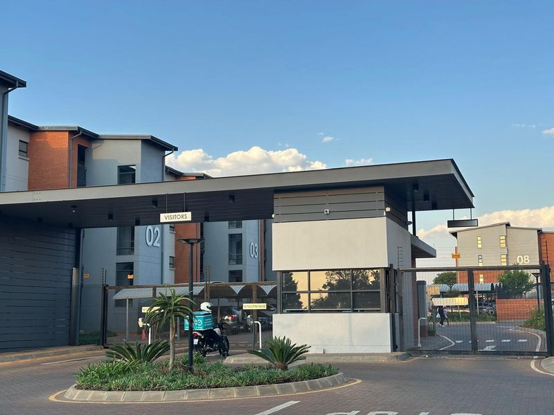 The main entrance to the apartment complex featuring a modern security gatehouse, automated access control, and well-maintained paved driveway.