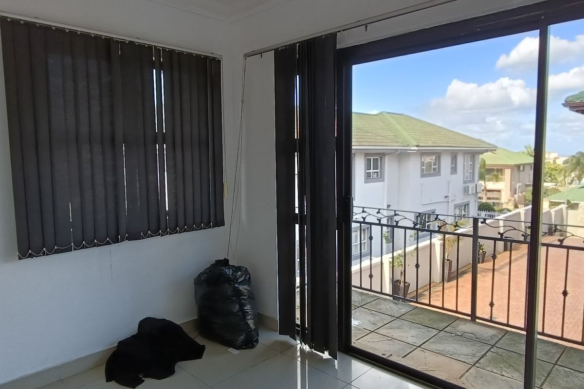 A bright, empty living area featuring tiled floors, vertical blinds, and a large sliding glass door providing access to a balcony.