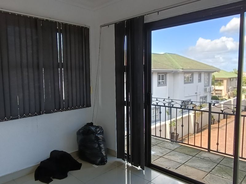 A bright, empty living area featuring tiled floors, vertical blinds, and a large sliding glass door providing access to a balcony.