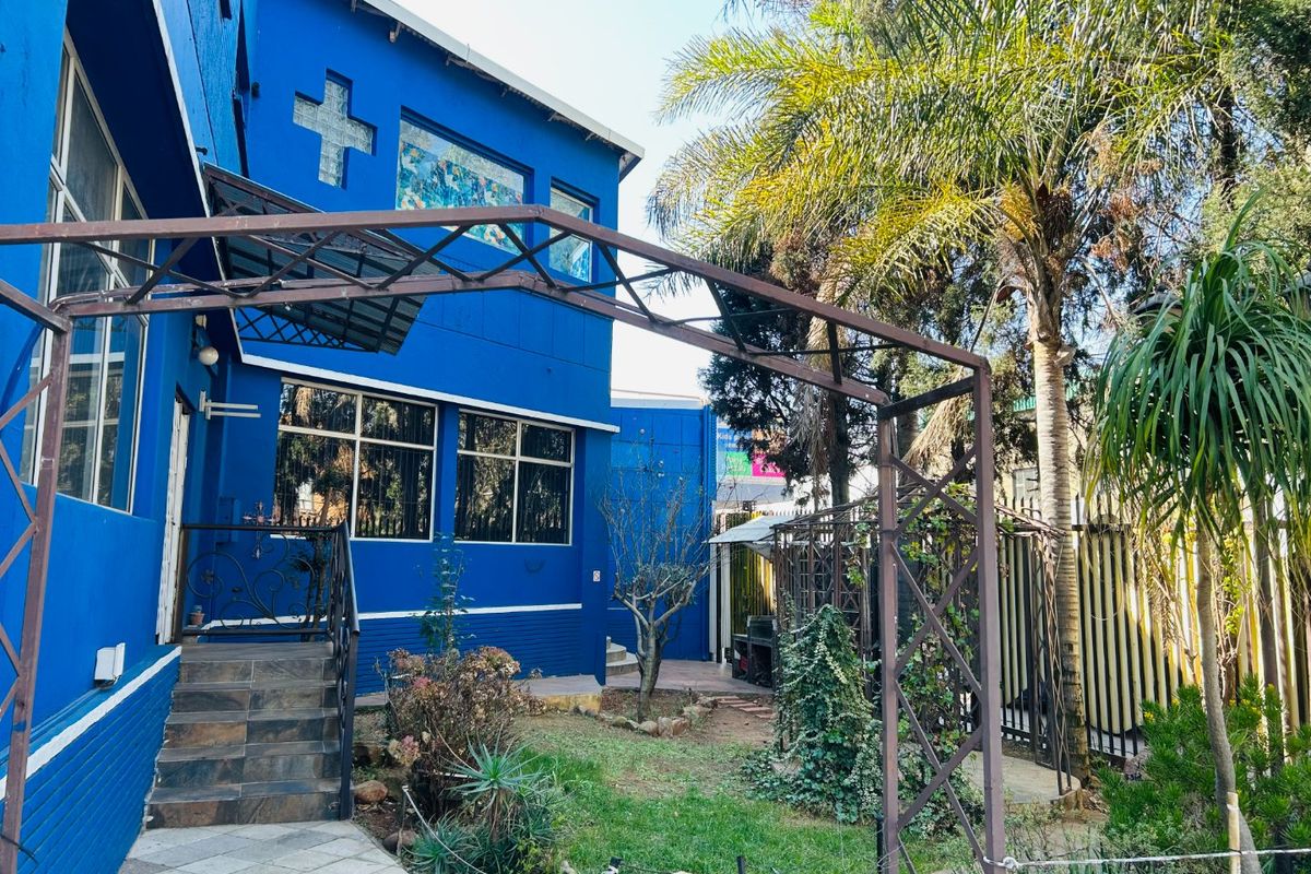 Rear exterior view of the commercial property featuring a blue facade, stained glass detailing, metal pergola, and a landscaped garden area with tiled steps.