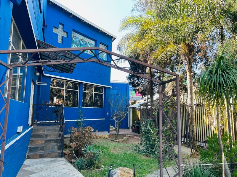 Rear exterior view of the commercial property featuring a blue facade, stained glass detailing, metal pergola, and a landscaped garden area with tiled steps.