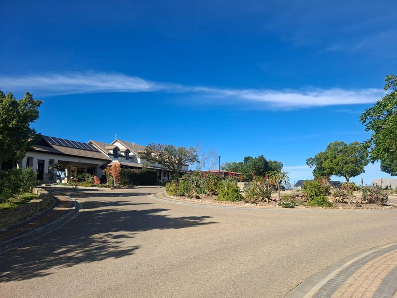 A well-maintained residential street view featuring a paved driveway, landscaped garden beds, and a building with roof-mounted solar panels under a clear sky.